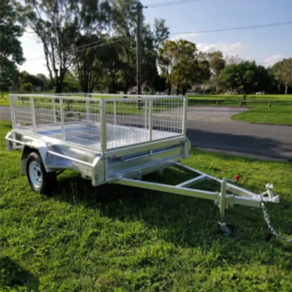 A 6x4 Single Axle Box Trailer ATM 750KG with mesh sides and a jockey wheel is parked on grass. It has two wheels, a tow hitch in the foreground, trees, and a paved road visible in the background.