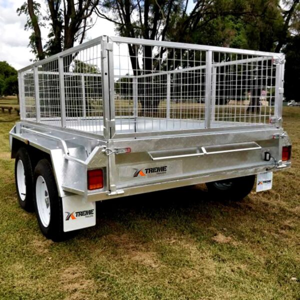 A Premium 8x6 Dual Axle Heavy Duty Tandem Trailer - 2000KG ATM with a silver frame and mesh sides is parked on grass, featuring rear lights and branded mudguards, while trees and a picnic bench are in the background.