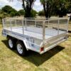 A Premium 8x6 Dual Axle Heavy Duty Tandem Trailer - 2000KG ATM with wire mesh sides and two red rear reflectors is parked on grass. Trees and a park setting are visible in the background beneath a partly cloudy sky.