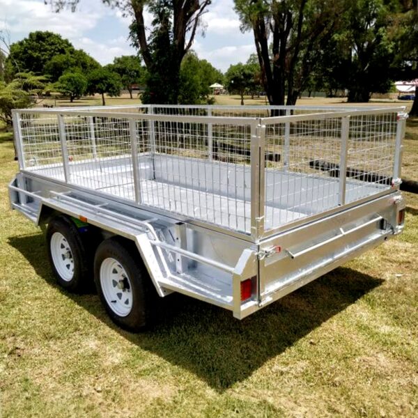 A Premium 8x6 Dual Axle Heavy Duty Tandem Trailer - 2000KG ATM with wire mesh sides and two red rear reflectors is parked on grass. Trees and a park setting are visible in the background beneath a partly cloudy sky.