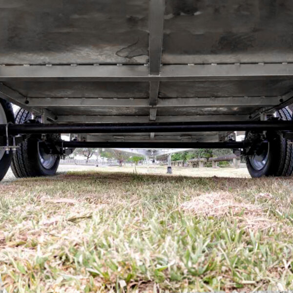 Ground-level view under the Premium 8x5 Dual Axle Heavy Duty Tandem Trailer (3200KG ATM), showcasing its wheels and axles while parked on grass, with trees and an open field in the background.