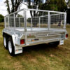 A Premium 8x5 Dual Axle Heavy Duty Tandem Trailer with a metal mesh enclosure is parked on grassy ground. It features a silver finish and branded sides, with trees and a picnic table in the background.