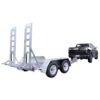 A black truck is towing a metallic utility trailer with dual axles and two large ramps at the back, designed for transporting heavy equipment or vehicles. The trailer is empty and parked on a plain white background.