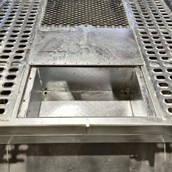 Industrial metal platform with a rectangular cutout in the center. The surface has perforated grids and a smooth section leading to an open compartment. The construction appears sturdy, with metallic textures prominent.