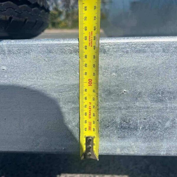 A yellow measuring tape hangs vertically over a 10x6 Galvanized Plant Trailer, showing measurements in centimeters and inches. A large tire is partially visible in the upper left corner, casting a shadow on the trailers metallic surface.