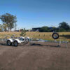 A 6.2m galvanized boat trailer with a 2000kg ATM for boats 5.7m-6.2m, featuring dual axles, a mounted spare tire, and multiple rollers, is parked on a paved area with a grassy field and trees in the background under a clear blue sky.