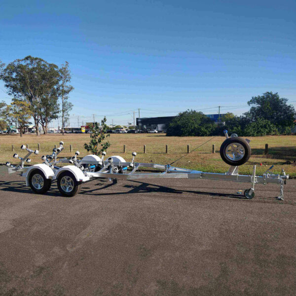 A 6.2m galvanized boat trailer with a 2000kg ATM for boats 5.7m-6.2m, featuring dual axles, a mounted spare tire, and multiple rollers, is parked on a paved area with a grassy field and trees in the background under a clear blue sky.