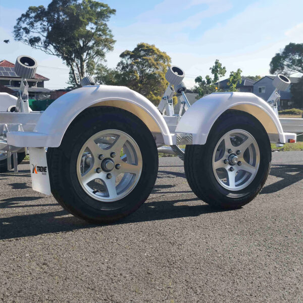 A close-up view of a 6.2m Galvanized Boat Trailer with tandem wheels and fenders parked on asphalt, designed for boats 5.7m-6.2m (18.7-20.3ft). Trees and houses blur in the background under a clear blue sky, while bright sunlight casts shadows on the ground.