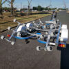 A 6.2m Galvanized Boat Trailer with 2000kg ATM, perfect for 5.7m-6.2m boats, features adjustable supports and wheels, decorated with white and blue elements plus reflectors. Its parked on a paved road near a park filled with trees and benches under a clear blue sky.