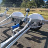 A 6.2m Galvanized Boat Trailer with 2000kg ATM, perfect for boats 5.7m-6.2m, is parked on pavement in a park. It features two large wheels and adjustable supports, with grass and trees under a clear blue sky in the background.