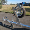 A close-up of a metallic hitch and mounted spare tire on the 6.2m Galvanized Boat Trailer (2000kg ATM) shows safety chains, a hand crank, and a blue wheel chock. The setup is on a paved surface with grass and trees in the background. Ideal for boats 5.7m-6.2m (18.7-20.3ft).