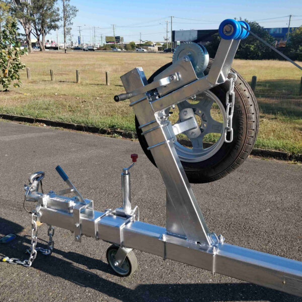 A close-up of a metallic hitch and mounted spare tire on the 6.2m Galvanized Boat Trailer (2000kg ATM) shows safety chains, a hand crank, and a blue wheel chock. The setup is on a paved surface with grass and trees in the background. Ideal for boats 5.7m-6.2m (18.7-20.3ft).