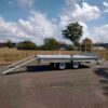 A Premium 12x7 Galvanized Flat Top Trailer with 3500KGS ATM and ramps, featuring a shiny metal surface and dual axles, is parked on a paved road against a backdrop of grass, trees, and a partly cloudy sky.