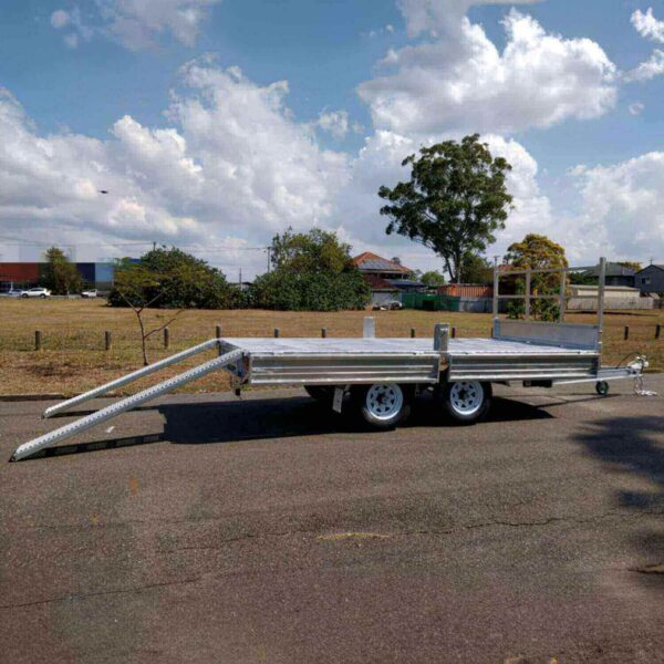 A Premium 12x7 Galvanized Flat Top Trailer with 3500KGS ATM and ramps, featuring a shiny metal surface and dual axles, is parked on a paved road against a backdrop of grass, trees, and a partly cloudy sky.