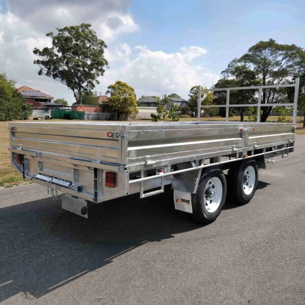 A Premium 14x7 Galvanized Flat Top Trailer with 3500KGS ATM and ramps, featuring two axles and side rails, is parked on a paved road against a backdrop of grass, trees, houses, and a partly cloudy sky.