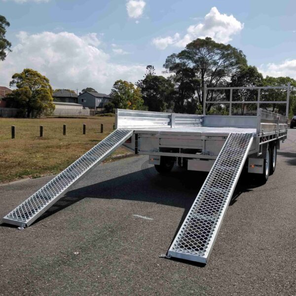 A Premium 10x7 Galvanized Flat Top Trailer with 3500KGS ATM and ramps sits on a paved road, near grass and trees under a partly cloudy sky. The trailer features metal railings on the front and sides.