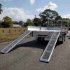 A Premium 14x7 Galvanized Flat Top Trailer with a 3500KGS ATM and two metal ramps is parked on a quiet suburban street lined with trees and grass, under a partly cloudy sky.