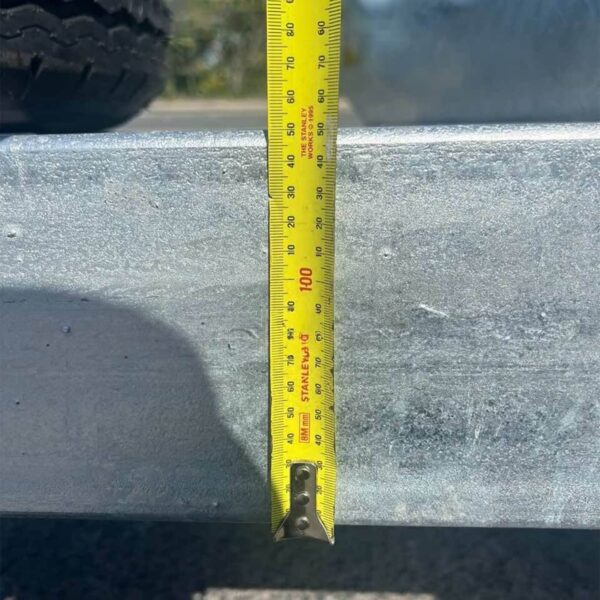 A yellow tape measure hangs vertically, measuring the height of a metal surface on the Premium 16x8 Galvanized Flat Top Trailer. The tape marks centimeters, showing 100 cm near the edge, with a wheel visible in the upper left corner.