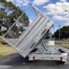 A 12×7 dual axle silver hydraulic 3-way tipper flat top trailer with a 3500KG ATM and ramps is raised at an angle, displaying its metal frame and mesh sides. Its parked on a tree-lined street under a partly cloudy sky.