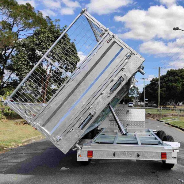 A 12×7 dual axle silver hydraulic 3-way tipper flat top trailer with a 3500KG ATM and ramps is raised at an angle, displaying its metal frame and mesh sides. Its parked on a tree-lined street under a partly cloudy sky.