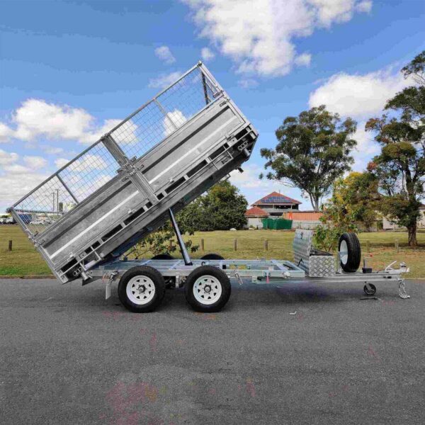A 12×7 Dual Axle Hydraulic 3-Way Tipper Flat Top Trailer - 3500KG ATM with Ramps is tilted upward on a road. It features dual wheels, a wire mesh gate, and an attached spare tire. The background reveals grass, trees, a house, and a blue sky with clouds.