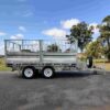 A 12×7 Dual Axle Hydraulic 3-Way Tipper Flat Top Trailer - 3500KG ATM is parked on asphalt. It includes mesh side panels and a spare tire on the front right. Trees and a partly cloudy blue sky fill the background.