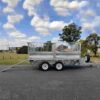 A silver 12×7 Dual Axle Hydraulic 3-Way Tipper Trailer - 3500KG ATM with extended ramps, featuring a mesh fence, is parked on a paved surface in front of trees and grass under a partly cloudy sky.