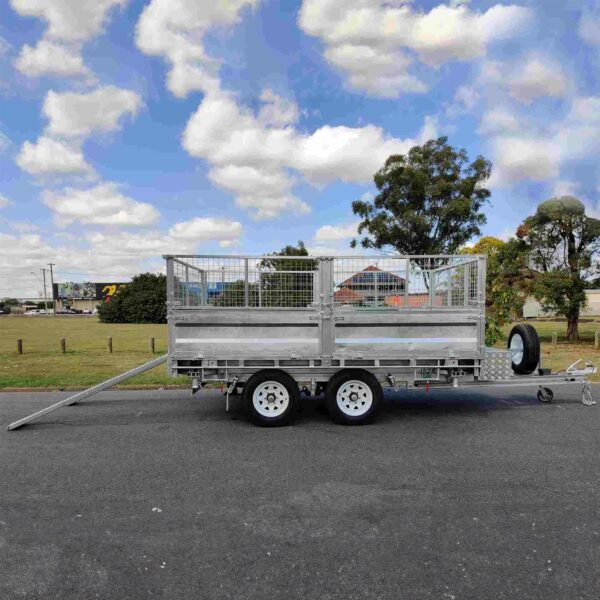 A silver 12×7 Dual Axle Hydraulic 3-Way Tipper Trailer - 3500KG ATM with extended ramps, featuring a mesh fence, is parked on a paved surface in front of trees and grass under a partly cloudy sky.