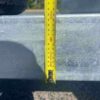 A 12×7 Dual Axle Hydraulic 3-Way Tipper Trailer is parked, its tape measure hanging from the sunlit metal surface shows around 90 cm in length. The trailer stands on a shadowed paved ground.