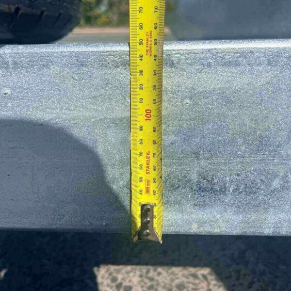 A 12×7 Dual Axle Hydraulic 3-Way Tipper Trailer is parked, its tape measure hanging from the sunlit metal surface shows around 90 cm in length. The trailer stands on a shadowed paved ground.