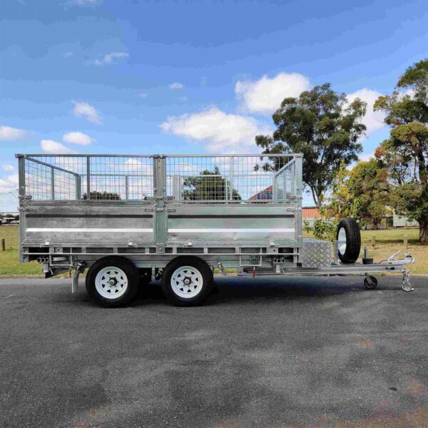 A 14×7 Dual Axle Hydraulic 3-Way Tipper Flat Top Trailer with a metal mesh cage, white wheels, and a front-mounted spare tire is parked on a paved surface, surrounded by trees under a clear blue sky.
