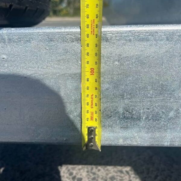 A measuring tape is vertically positioned against a metallic surface of the 14×7 Dual Axle Hydraulic 3-Way Tipper Flat Top Trailer - 3500KG ATM with Ramps, reading just over 90 centimeters. A shadow is cast on the textured ground in the background.