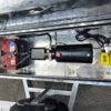 The 12×7 Dual Axle Hydraulic Tipper Tandem Box Trailer - 3500KG ATM With Ramps features a metal compartment with a large battery, a black cylindrical device with wires, and a small yellow control box, all mounted on its textured metallic surface.
