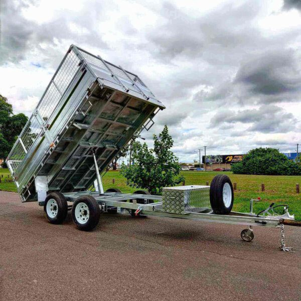 A 12×7 Dual Axle Hydraulic Tipper Tandem Box Trailer - 3500KG ATM With Ramps, featuring a raised bed and mounted spare tire, stands on asphalt under cloudy skies with trees and a building in the backdrop.
