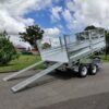 A 12×7 Dual Axle Hydraulic Tipper Tandem Box Trailer - 3500KG ATM with ramps, featuring metal mesh side panels, is parked on asphalt. In the background are green grass, trees, and a cloudy sky.