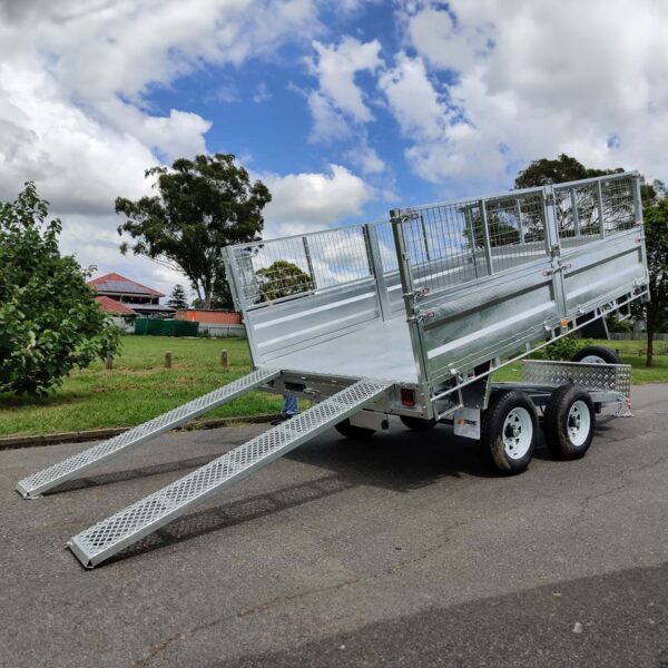 A 12×7 Dual Axle Hydraulic Tipper Tandem Box Trailer - 3500KG ATM with ramps, featuring metal mesh side panels, is parked on asphalt. In the background are green grass, trees, and a cloudy sky.