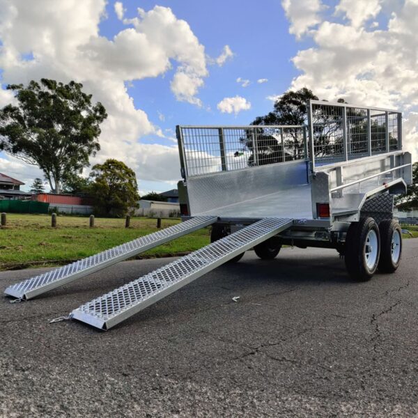 A 10×5 Dual Axle Hydraulic Tipper Tandem Box Trailer - 3500KG ATM with mesh sides is parked on a paved surface, featuring two loading ramps extended to the ground. Trees and clouds fill the background.