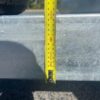 A 10×5 Dual Axle Hydraulic Tipper Tandem Box Trailer - 3500KG ATM With Ramp Holders is placed on a rough gray surface as a yellow measuring tape held vertically against its metal part shows about 90 centimeters.