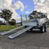 The 10×6 Dual Axle Hydraulic Tipper Tandem Box Trailer - 3500KG ATM With Ramp Holders, featuring a grated surface and dual axles, is shown parked on pavement. Two ramps extend from the back, with green grass and trees visible under a cloudy sky in the background.