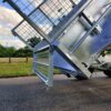 Close-up of the 10×6 Dual Axle Hydraulic Tipper Tandem Box Trailer - 3500KG ATM With Ramp Holders, featuring a raised mesh gate, parked on a paved road. The background showcases grass, trees, and a partly cloudy sky.