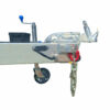 Close-up of a 12x7 Dual Axle Flat Top Cattle/Livestock Trailers hitch assembly, with a metal coupler, safety chain, support wheel, and hand crank. It connects to a rectangular frame with side rails and ramps; a black component is visible in the background.