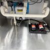 The 3.8m Big Box Food Trailer features under-sink plumbing with pipes, a water filtration system, two pumps, a tank with a caution sign on a metal base, and electrical wiring.