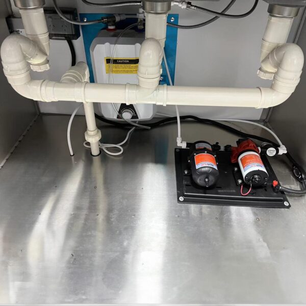 The 3.8m Big Box Food Trailer features under-sink plumbing with pipes, a water filtration system, two pumps, a tank with a caution sign on a metal base, and electrical wiring.