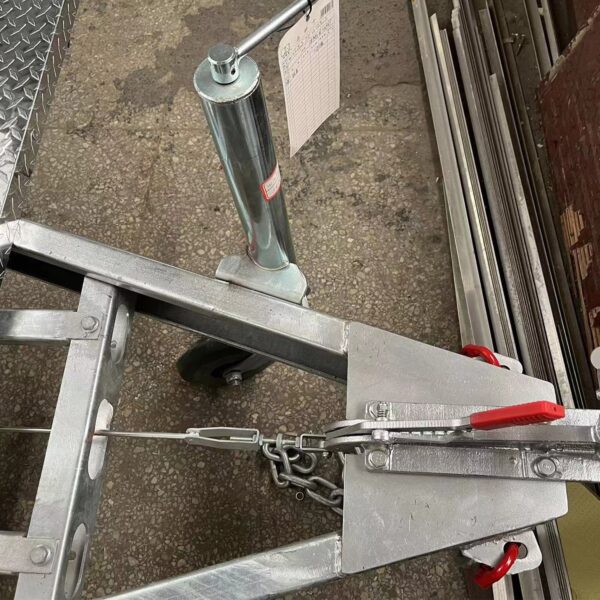 Close-up of a metal hitch on the 3.8m Big Box Food Trailer positioned on a textured concrete floor, featuring a red handle, chain, and support wheel. Nearby are metal rods and a clipboard with paper.
