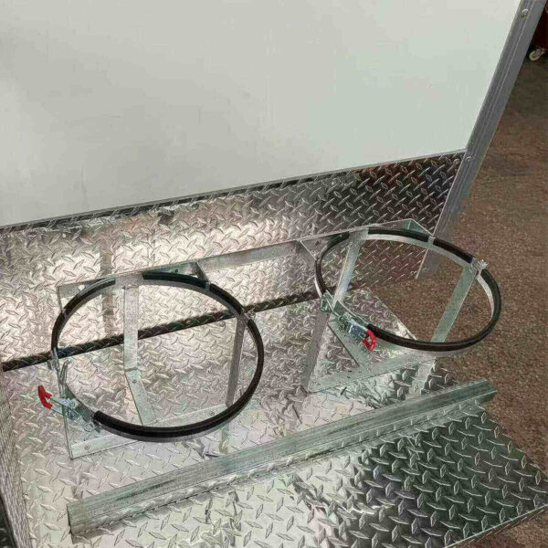 The 3.8m Big Box Food Trailer features a metal stand with two circular brackets suitable for tanks or drums. It sits on a diamond plate surface with a simple frame and clamps for secure holding, reflecting light for a shiny appearance.