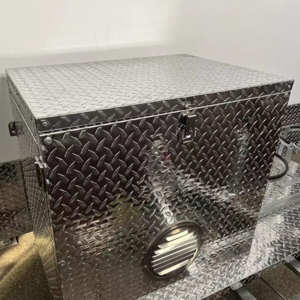 The 3.8m Big Box Food Trailer features a silver metallic exterior with a diamond plate pattern, secured by a latch and lock. Resting on its matching industrial surface, it boasts a side circular vent with horizontal slats.