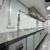 The 3.8m Big Box Food Trailer boasts a clean, modern kitchen with stainless steel countertops, white cabinetry, electrical outlets, and overhead storage. Its long and narrow space includes a non-slip patterned metal floor.