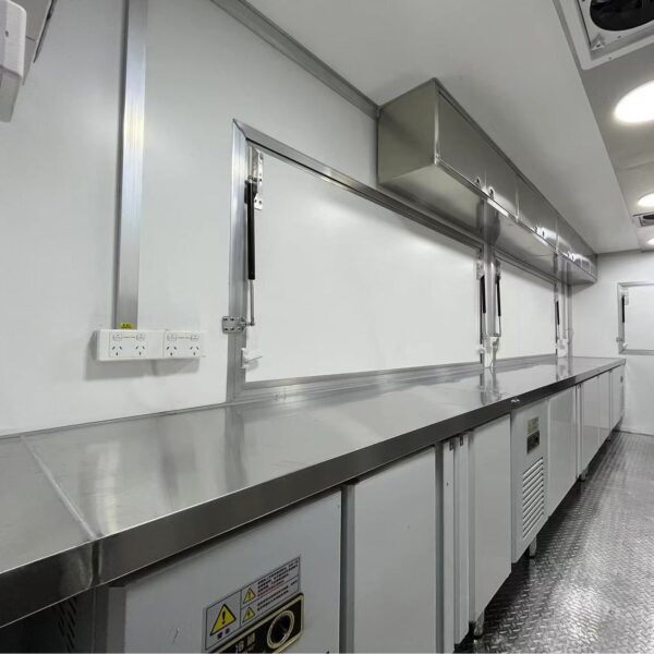 The 3.8m Big Box Food Trailer boasts a clean, modern kitchen with stainless steel countertops, white cabinetry, electrical outlets, and overhead storage. Its long and narrow space includes a non-slip patterned metal floor.