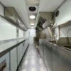 Inside the 3.8m Big Box Food Trailer, youll find stainless steel countertops, a sink, and overhead ventilation in a narrow space with a checkered metallic floor designed for optimal functionality and mobility.