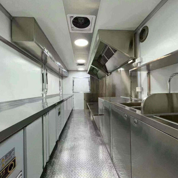 Inside the 3.8m Big Box Food Trailer, youll find stainless steel countertops, a sink, and overhead ventilation in a narrow space with a checkered metallic floor designed for optimal functionality and mobility.